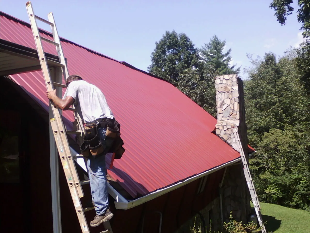 Roofer on a ladder beside a red metal roof and stone chimney — Soffit & Fascia Repair by Roofing Brothers in Salters