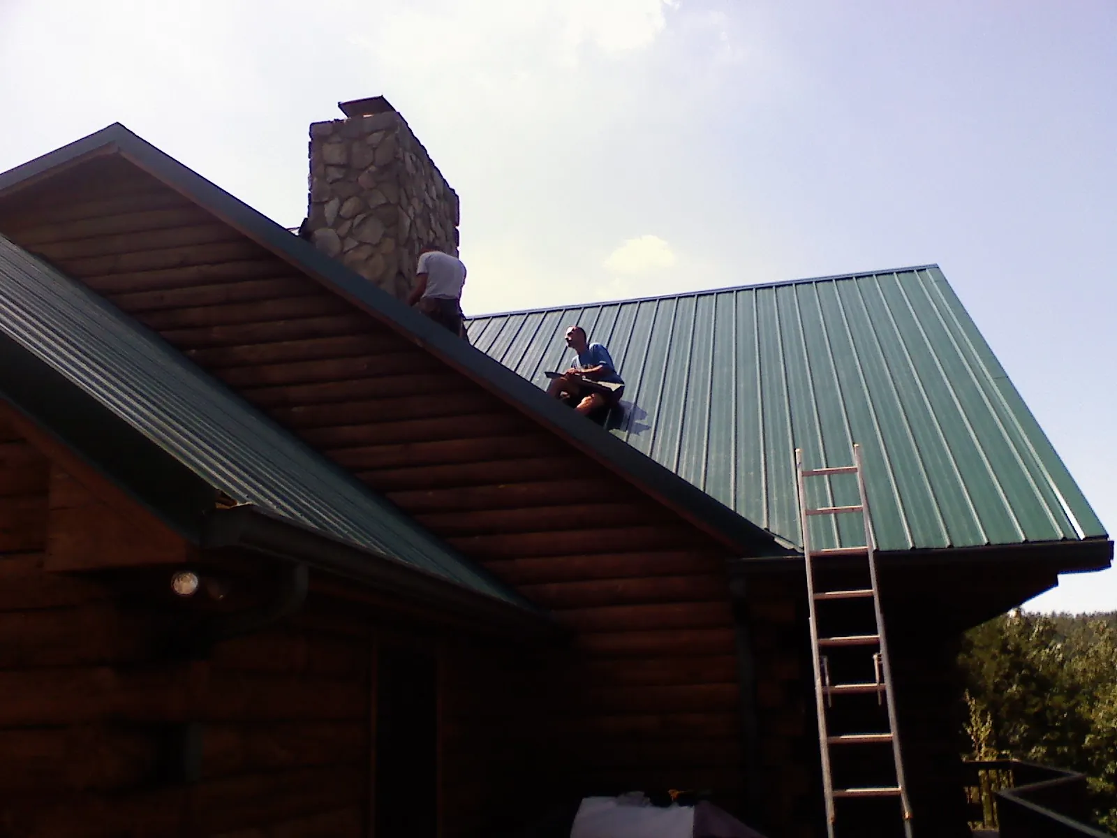 Roofers installing green metal panels beside a stone chimney on a log home in Salters